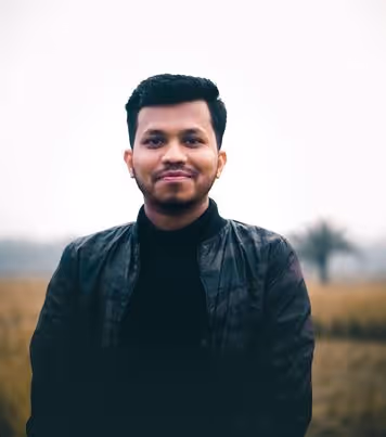 Smiling young man with short black hair wearing a black jacket standing outdoors with a blurred natural background.